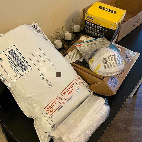 A pile of opened packages and respiratory N95 masks rests on a table, suggesting recent unpacking or sorting, in an indoor setting. The text on packaging includes "PRIORITY MAIL" and "STANLEY personal protection N95 Disposable Respirator."